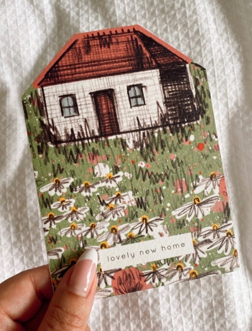 Card with a house and floral garden held by a hand on a white background.