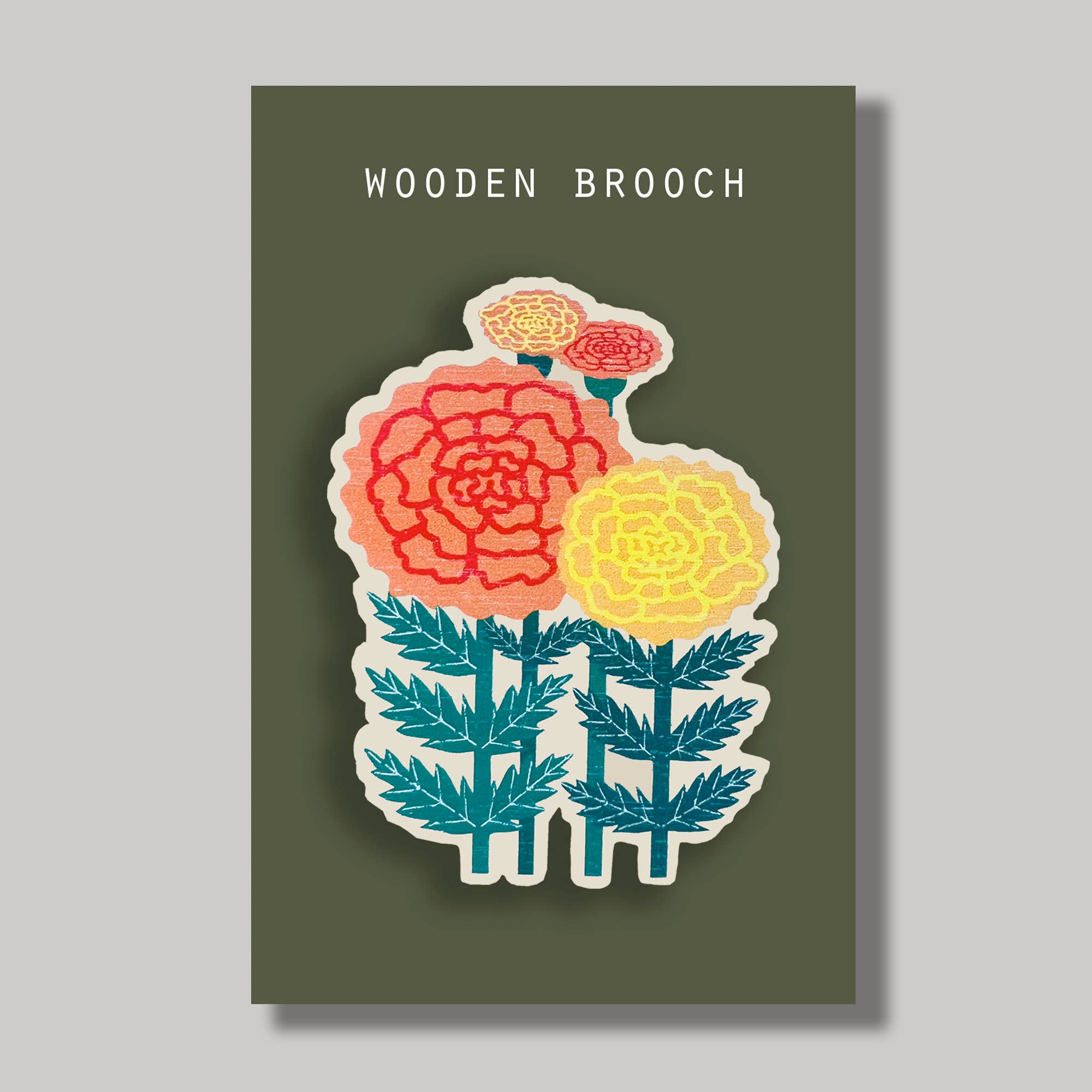 A wooden brooch featuring a stylised floral design with a large coral-pink flower, a yellow flower, teal leaves, and two smaller pink flowers on a dark olive green backing card. The backing card is labeled 'WOODEN BROOCH' in white text and is shown against a light grey background.
