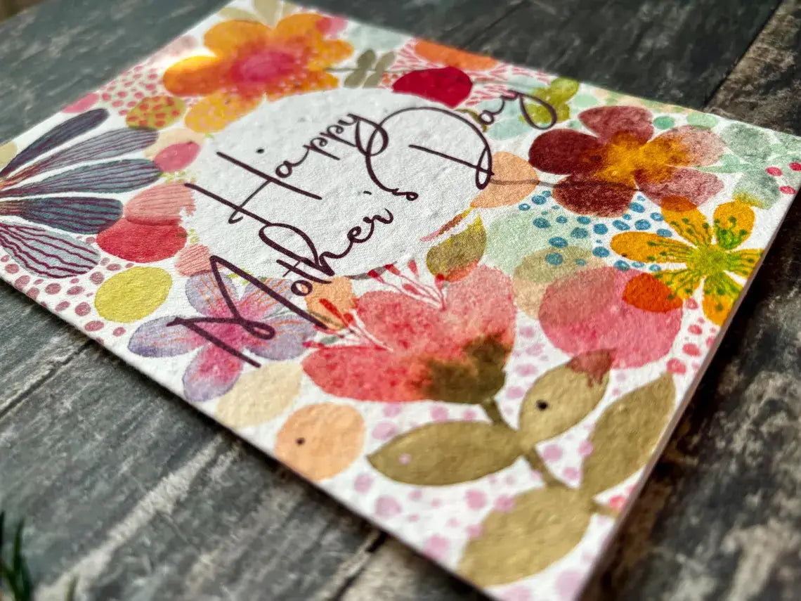 A Mother's Day greeting card with a colourful floral watercolor pattern and the words 'Happy Mother's Day' in black script, resting on a dark rustic wooden surface.