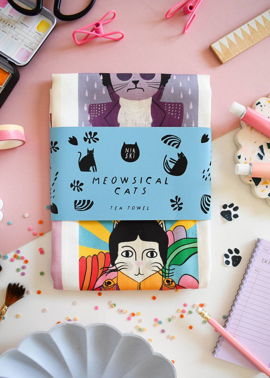 Two tea towels with cat designs on a pink surface with stationery items.