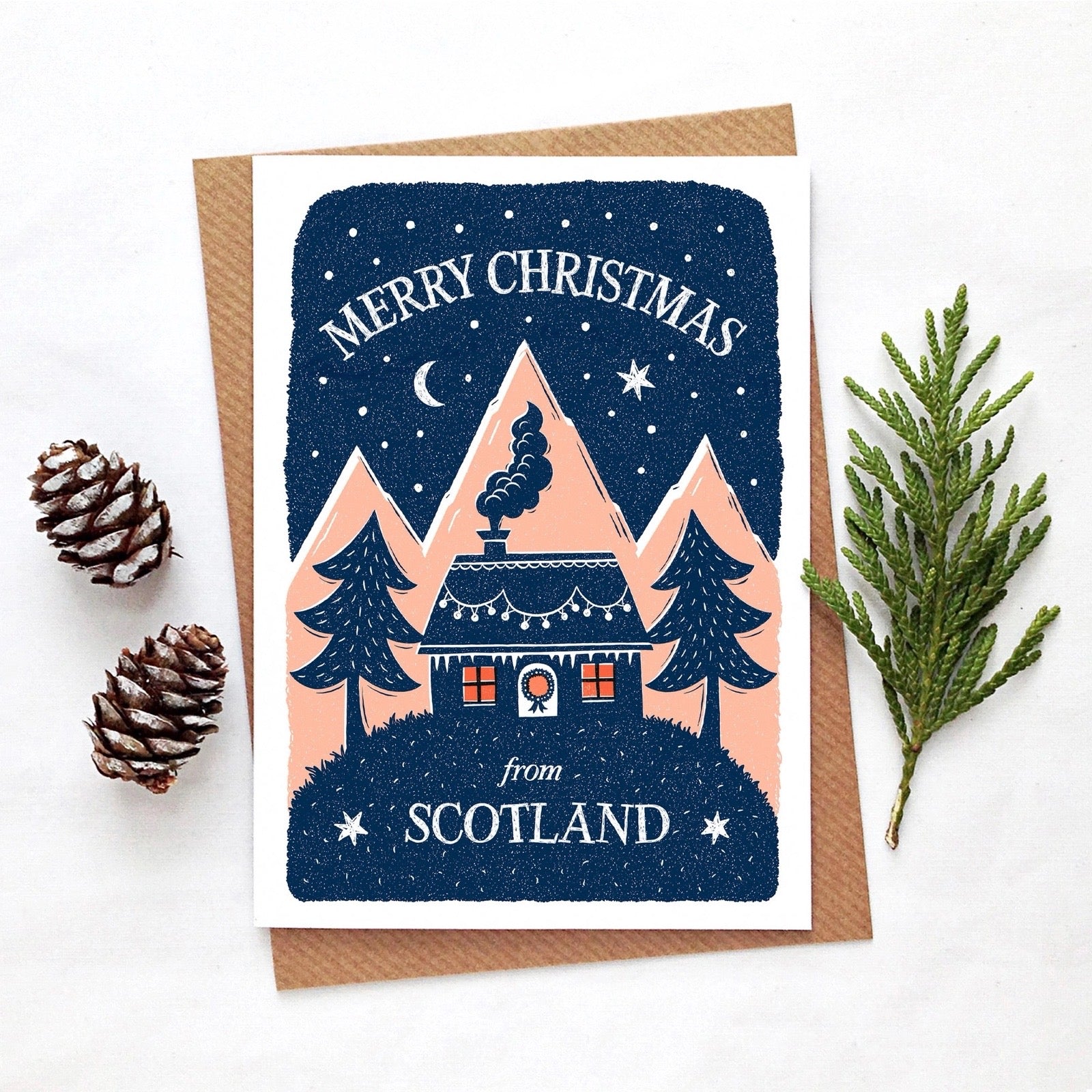 Christmas card with a house, trees, and 'Merry Christmas from Scotland' text on a blue background, sat on a table with pinecones and a branch.