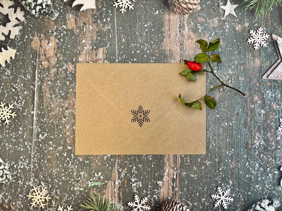 A brown kraft paper envelope with a printed black snowflake in the centre, placed on a rustic dark wooden surface decorated with white snowflakes, pinecones, evergreen branches, and a small red berry sprig.