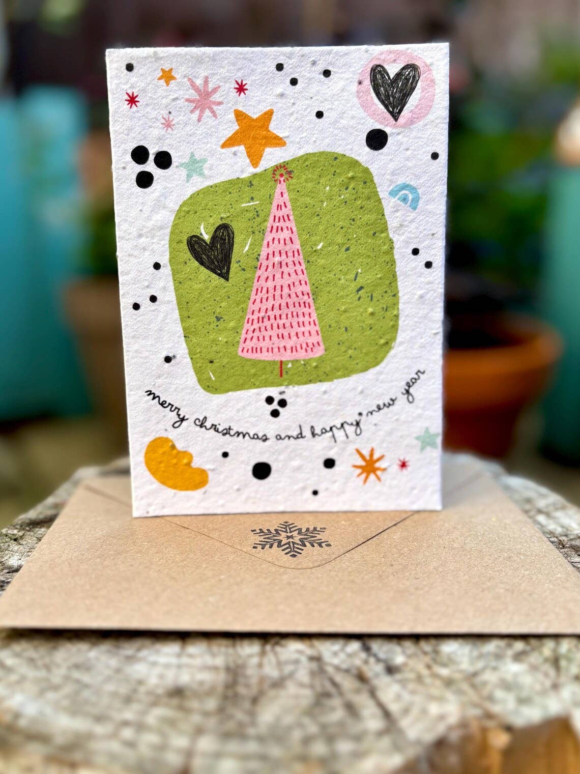 A handmade Christmas greeting card with an off-white background, featuring a green square, a pink Christmas tree, and hand-drawn black and orange accents. The card is displayed standing upright on a brown kraft envelope, set against a blurred outdoor background of potted plants.