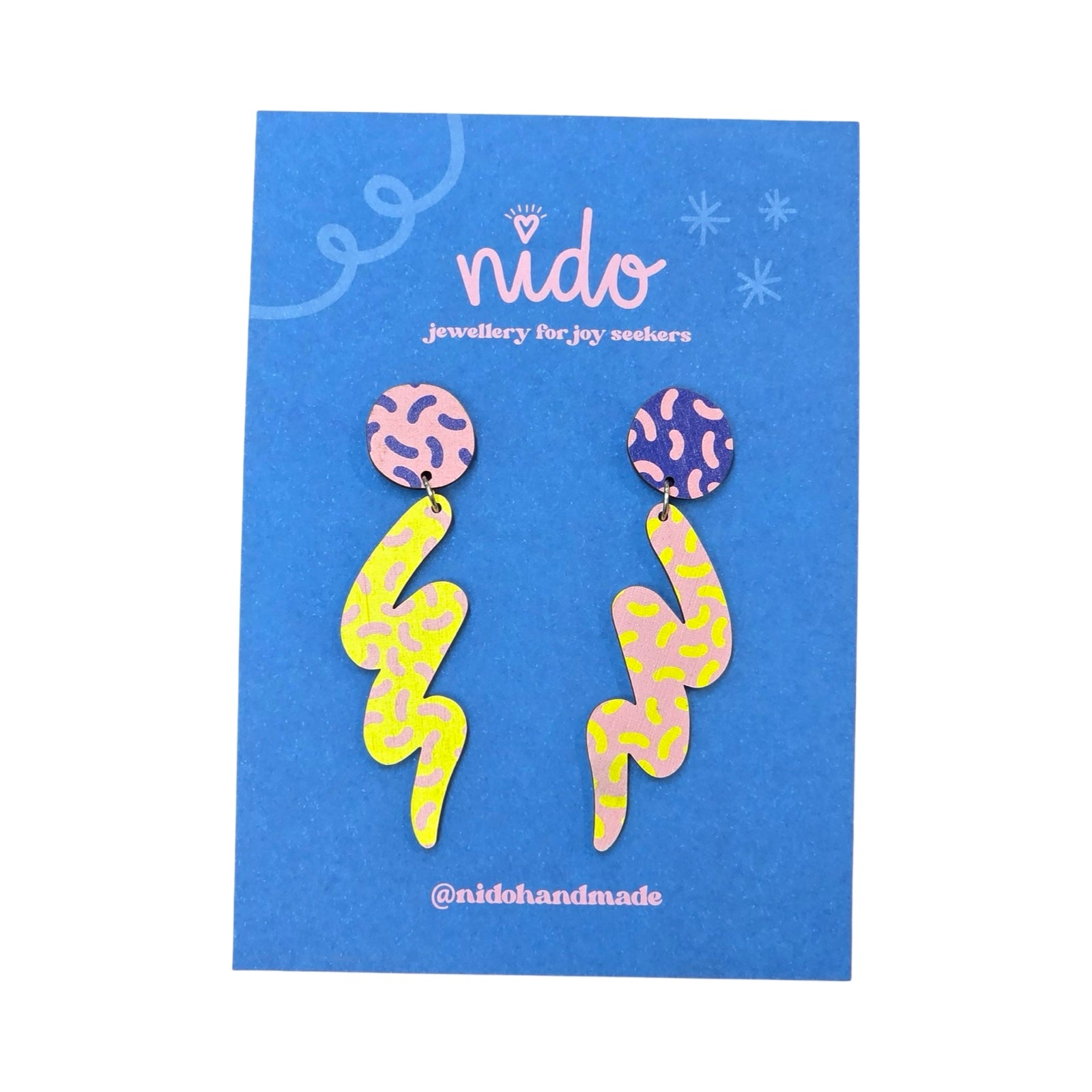 These wooden mismatched earrings feature one earring with a blue and pink dashed circular stud with a large pink and yellow dashed squiggle hanging underneath and one earring with a pink and blue dashed circular stud with a large yellow and pink dashed squiggle hanging underneath, presented on a branded blue backing card.