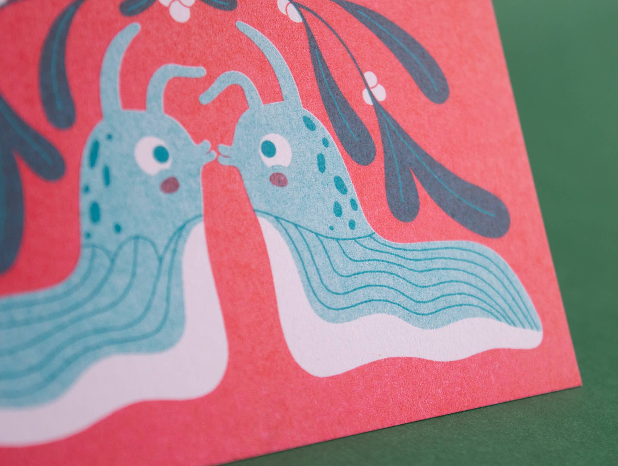 A close-up of a colourful card featuring two blue slugs facing each other on a coral-red background, with dark leaf-like shapes above and a green surface visible around the edges.