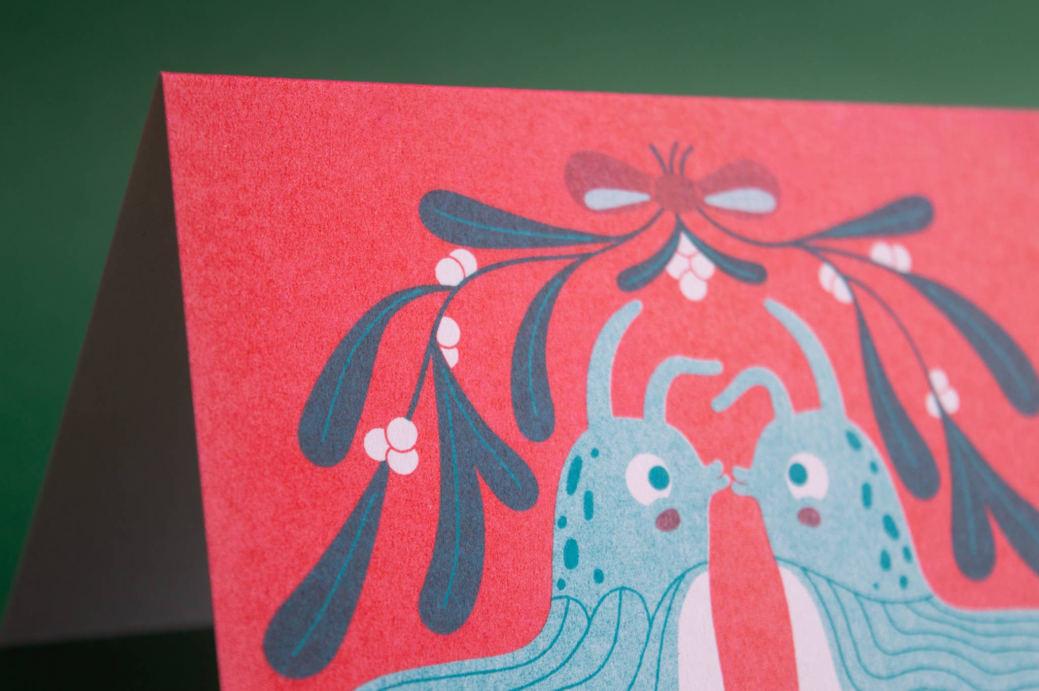 A close-up of a red greeting card featuring a whimsical illustration of two blue creatures under mistletoe, set against a dark green background.