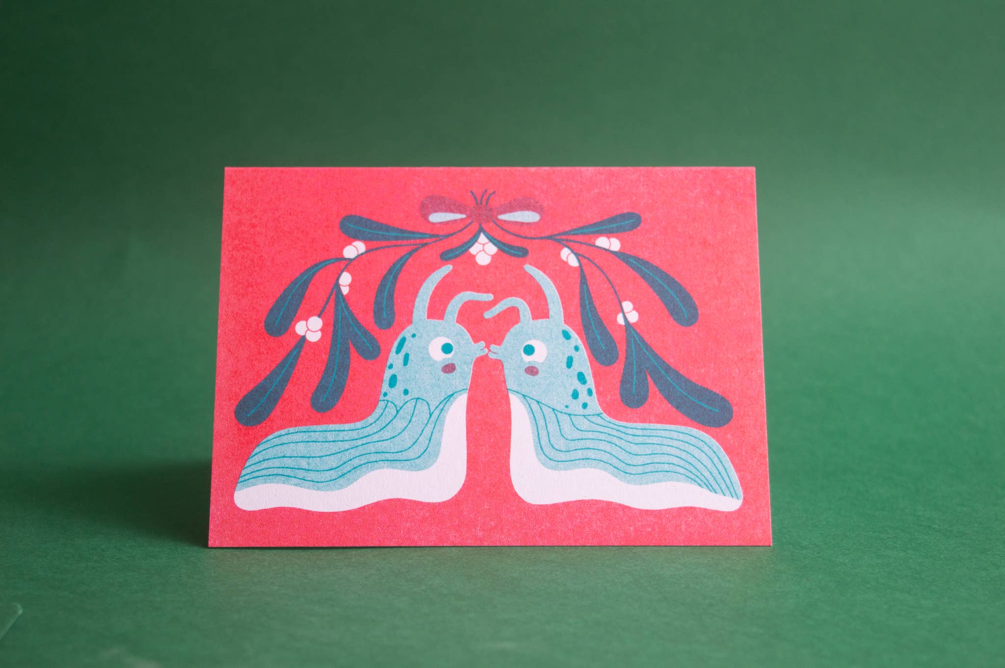 A red greeting card featuring a folk-art style illustration of two blue slugs facing each other beneath mistletoe, displayed on a solid green background.