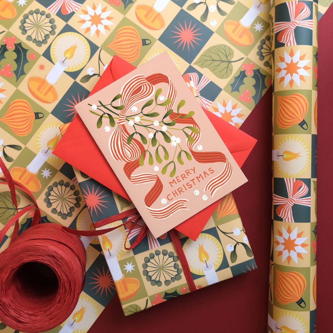 A Christmas card with mistletoe and a bow, sat on top of a gift wrapped present, with a roll of wrapping paper beside the gift.