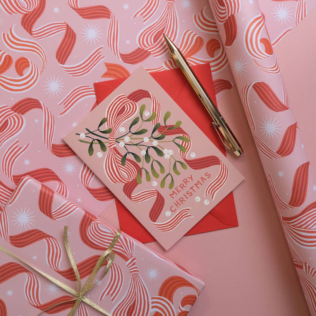 Pink Christmas gift wrapping paper with a red and orange bow pattern, shown wrapped around a gift box, a card, and a pen on a matching pink background.
