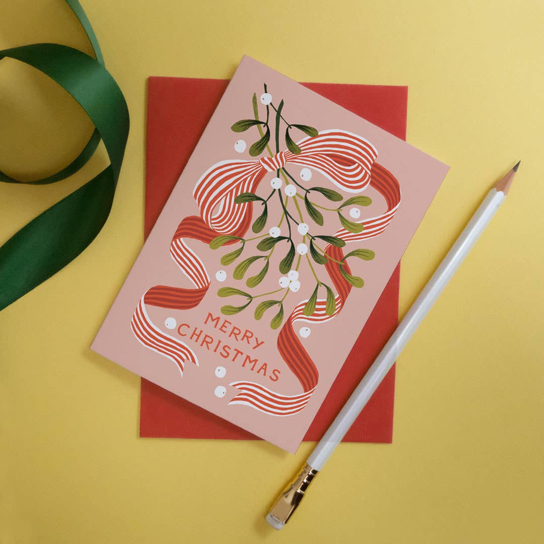 A pink Christmas greeting card with a red envelope, styled on a yellow background with a green ribbon and a white pencil.