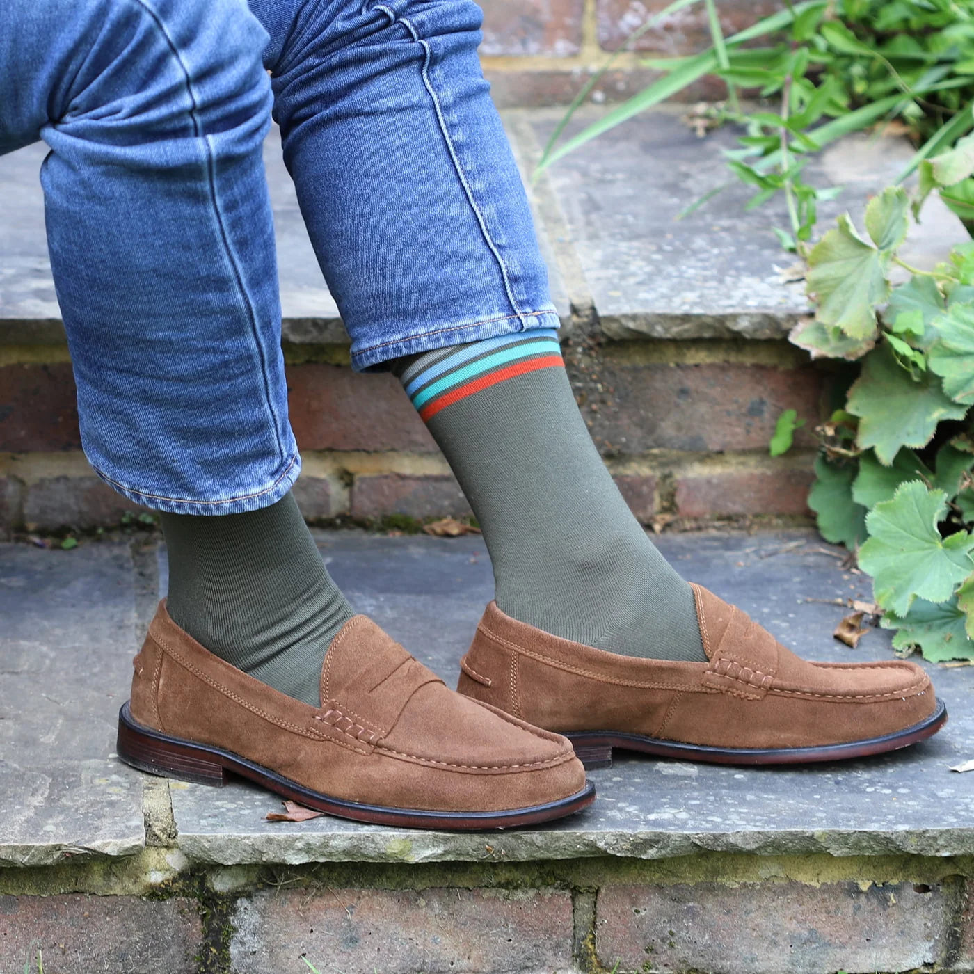 A person sitting on outdoor stone steps wearing brown suede penny loafers, olive green crew socks with thin red and teal stripes, and blue jeans.