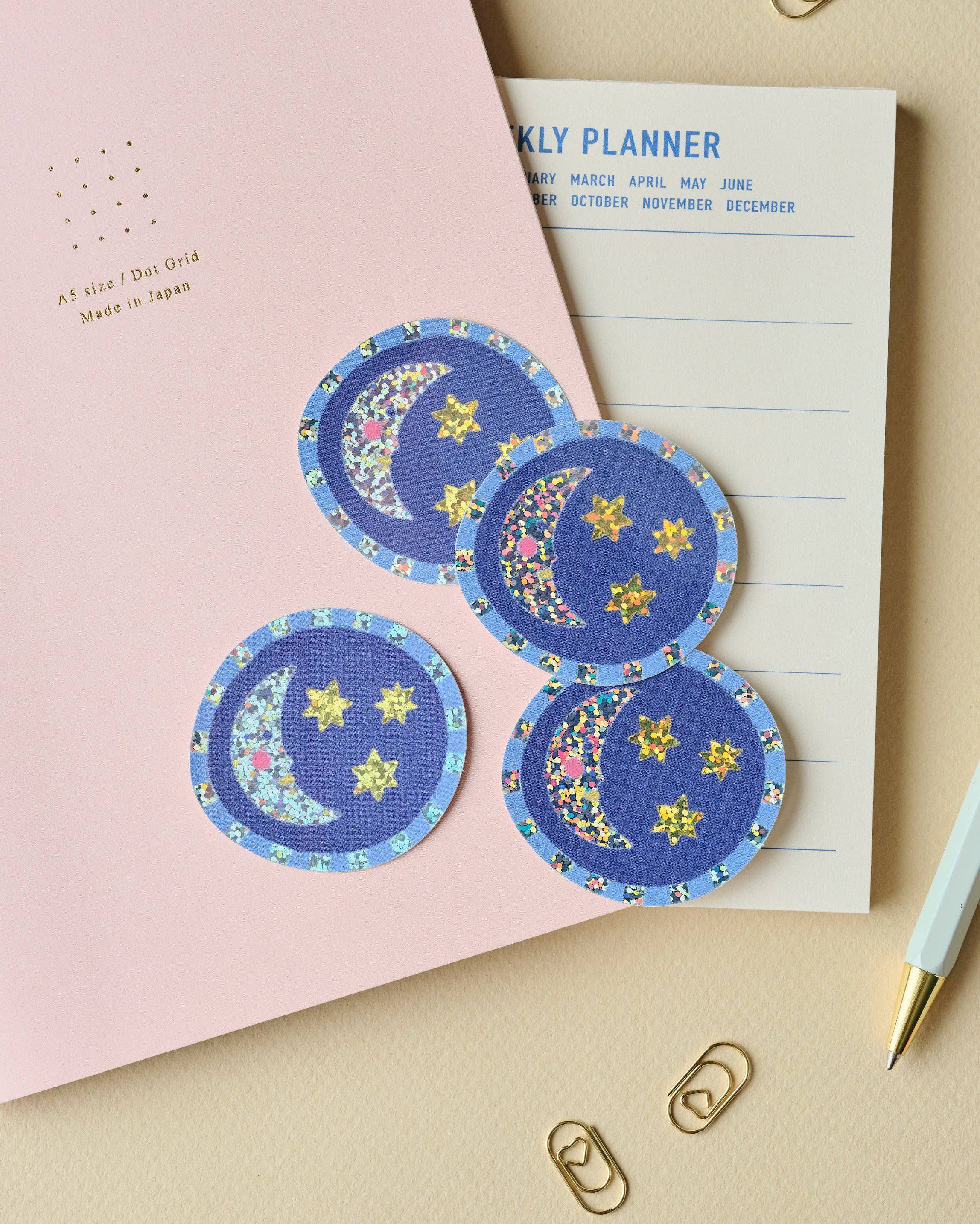 Round decorative stickers with blue celestial designs (crescent moons and stars) are arranged on a pink notebook and a white weekly planner pad, with gold paperclips on a beige surface.