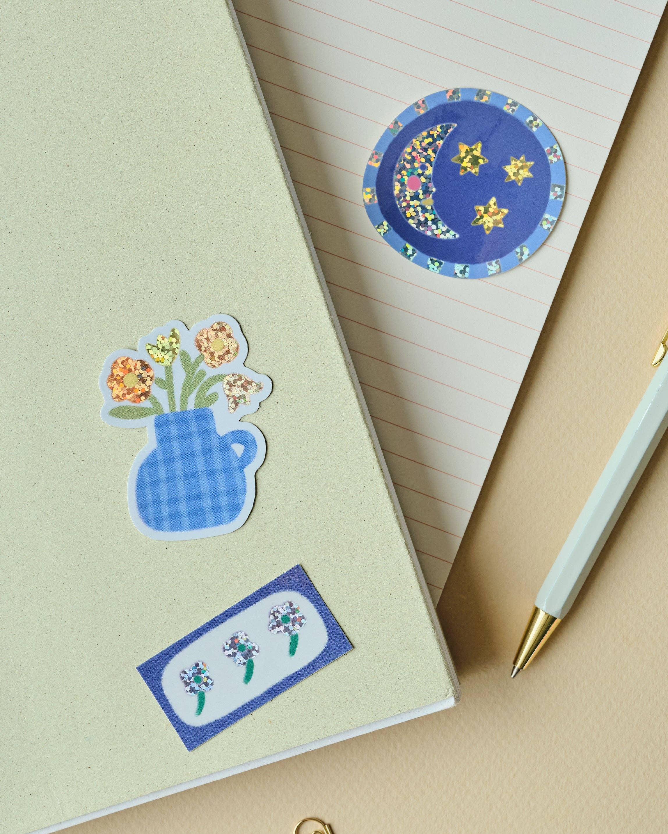 A styled flat-lay of a cream-coloured notebook with decorative stickers, including a blue gingham flower vase and a blue floral label, alongside a white pen with gold accents on a beige background.