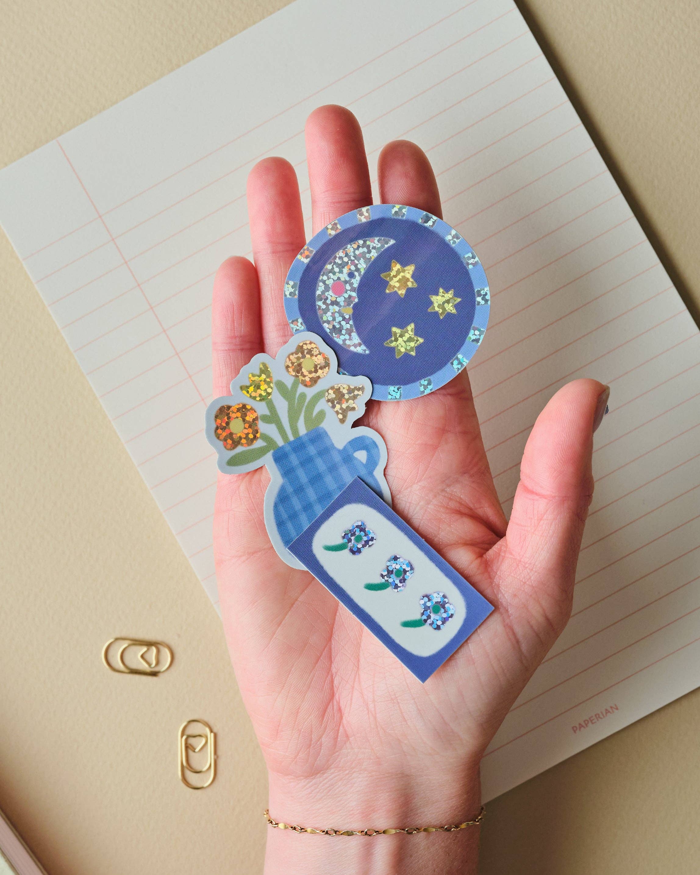 A hand holds three decorative stickers featuring a blue crescent moon and stars, small flowers, and a blue-and-white vase with floral details, placed over an open lined notebook.
