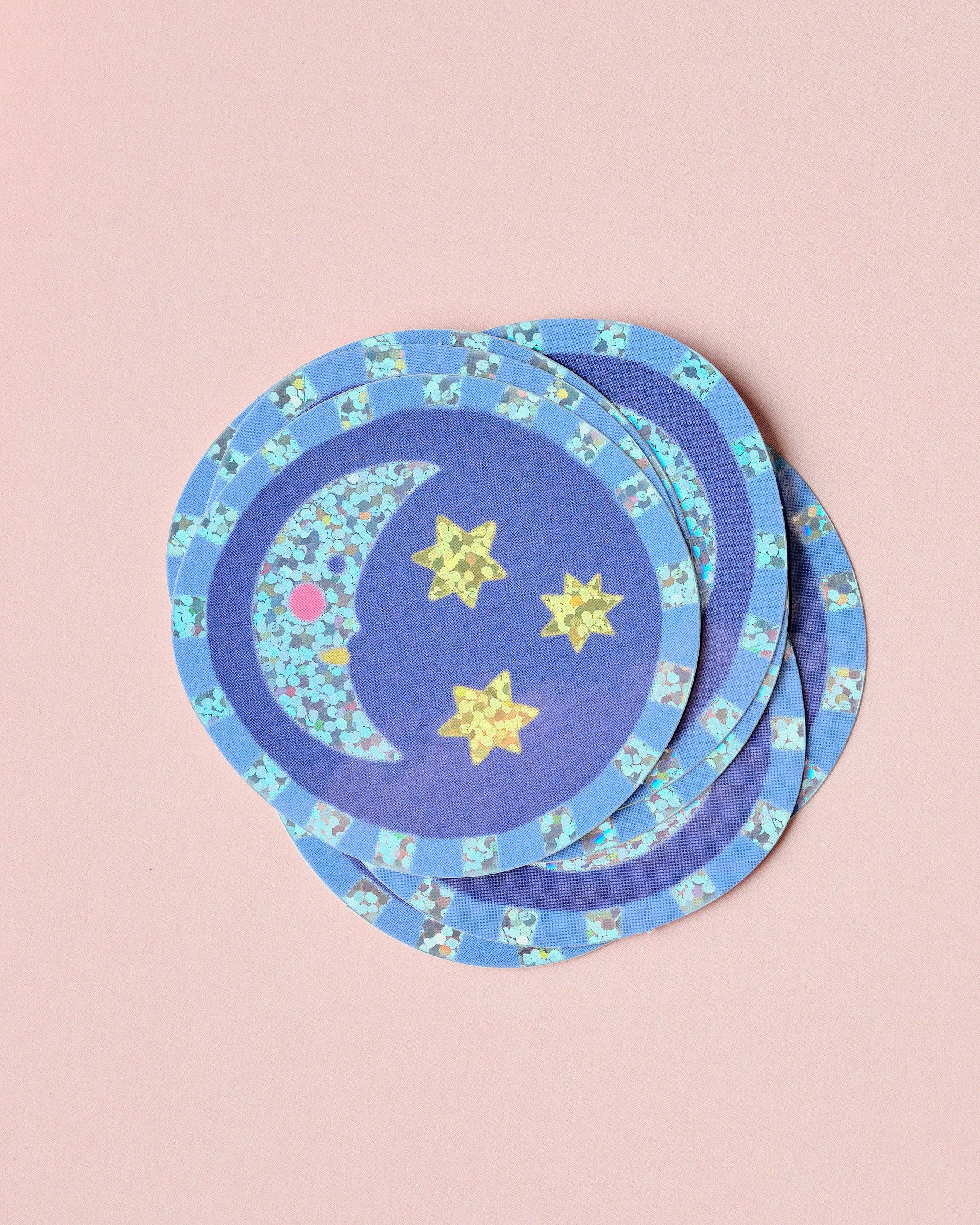A stack of round blue stickers with a celestial pattern featuring yellow stars, a crescent moon, and a patterned outer rim, displayed on a solid pink background.