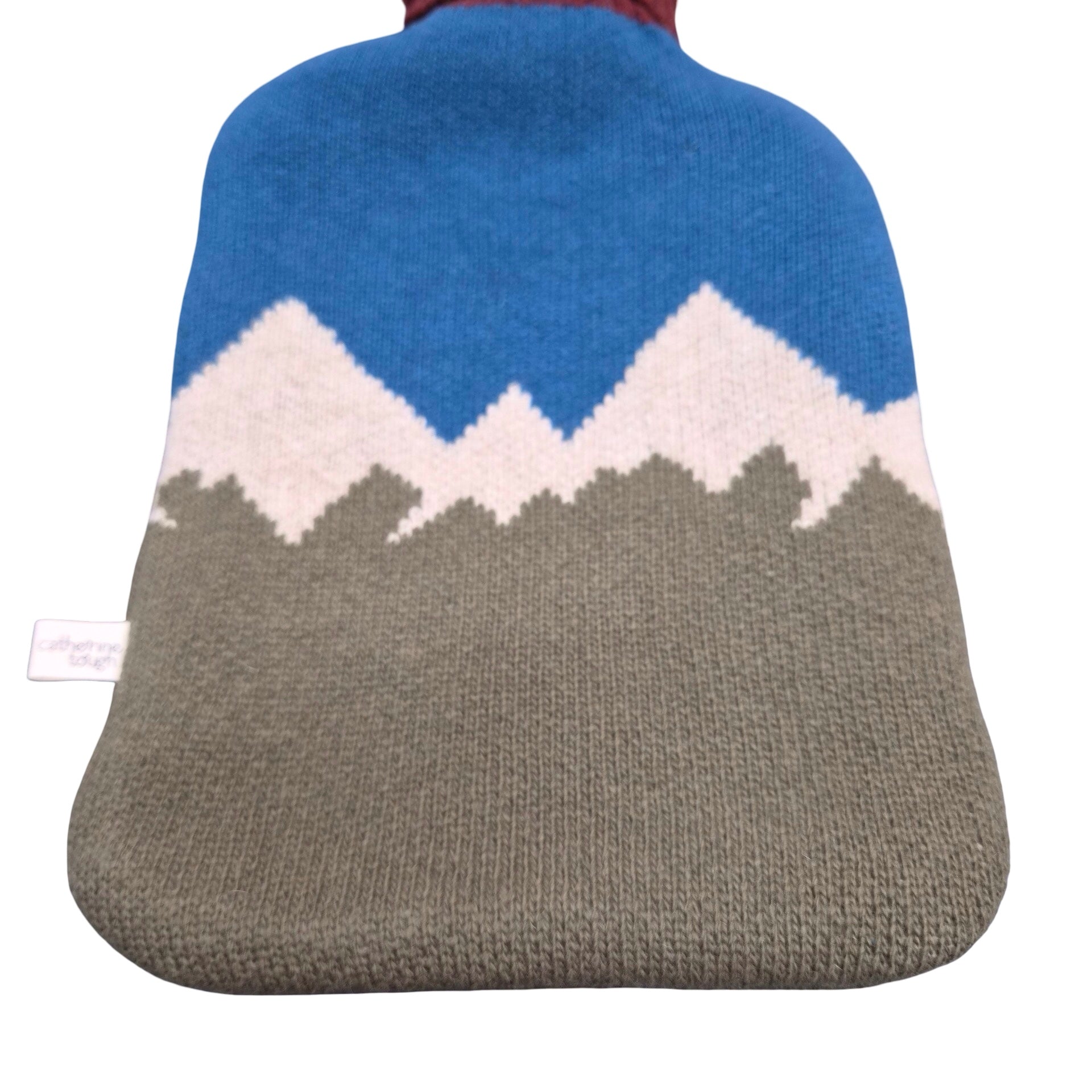 A knitted hot water bottle cover featuring a mountain pattern with a blue sky, white snow, and a grey foreground, shown against a plain white background.