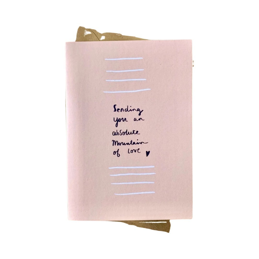 A sympathy card with a pale pink background, featuring the text 'SENDING YOU AN ABSOLUTE MOUNTAIN OF LOVE' in black, accompanied by a small black heart, and lined with a striped pattern.