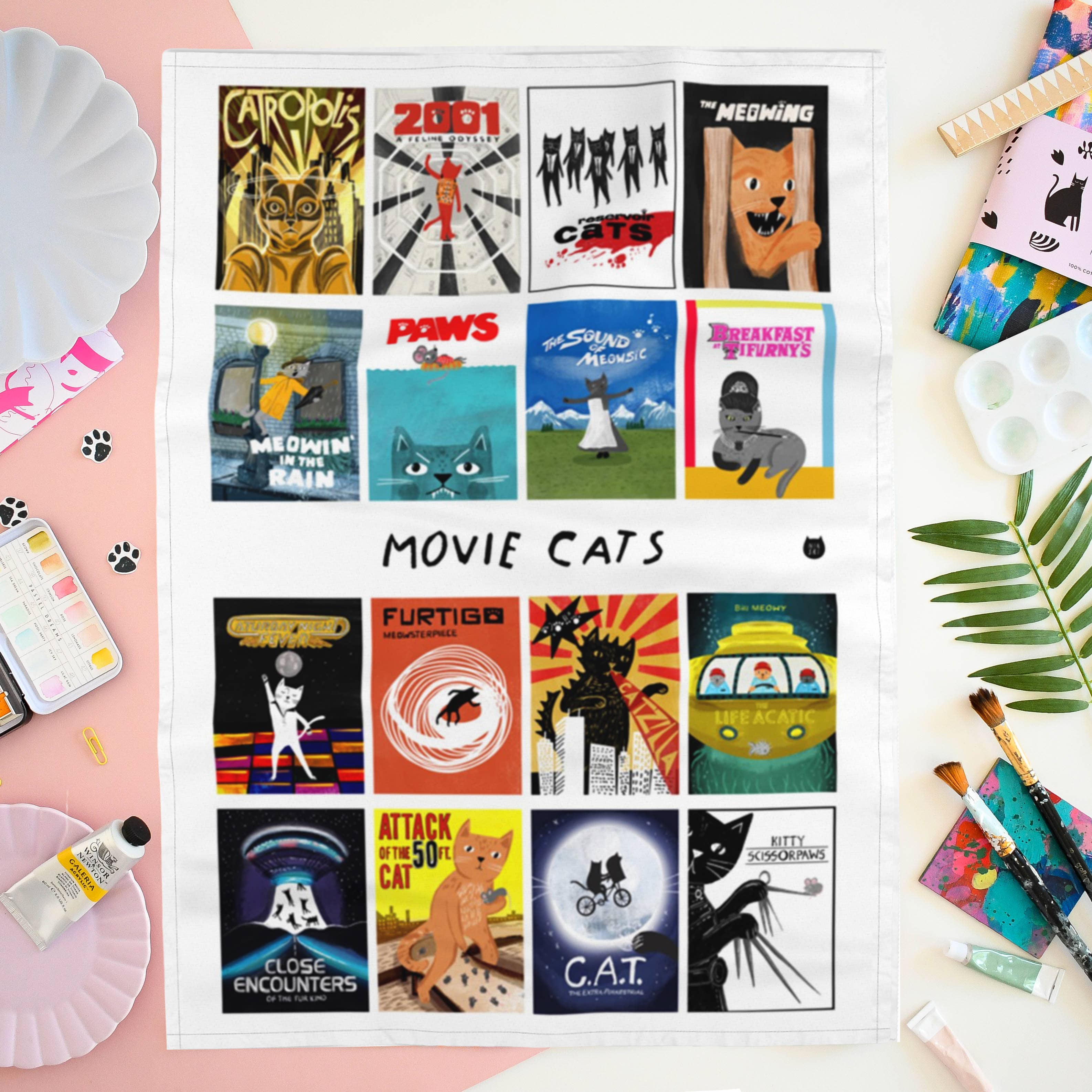 A white tea towel featuring a grid of 15 movie poster illustrations with the text 'MOVIE CATS' in the centre, on a pink background with art supplies and a palm leaf.