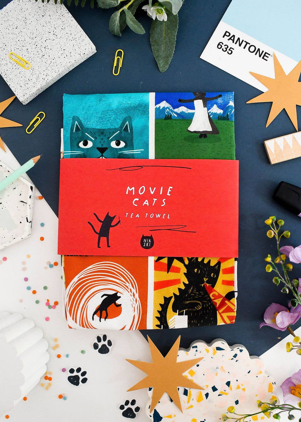 A tea towel packaged with a red band, on a table with art supplies, stationary and flowers.
