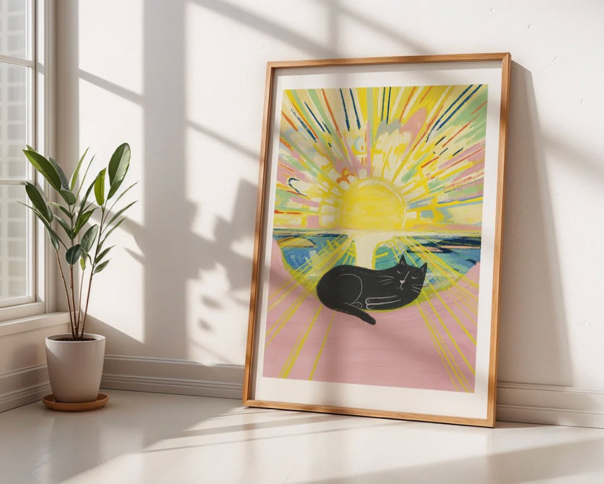 Framed abstract artwork with a black cat and sun on a pink background, placed on a light wooden floor with a plant nearby.
