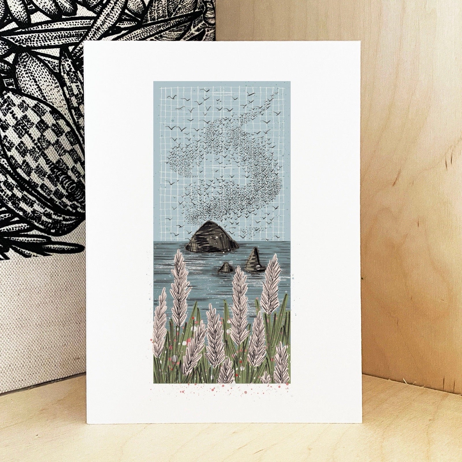 Framed artwork of a coastal scene with plants and rocks on a wooden surface.