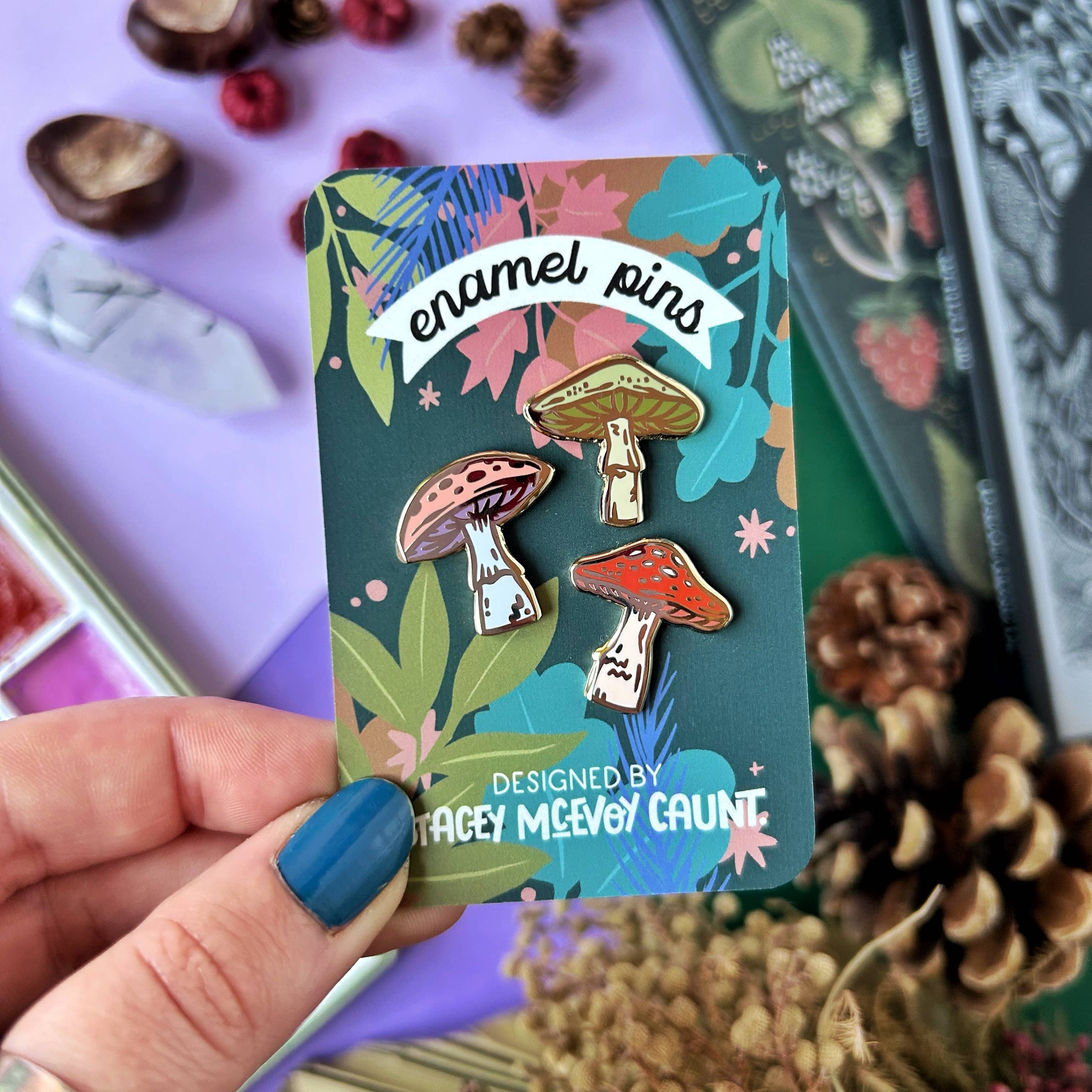 Card with mushroom-themed enamel pins held by a hand, surrounded by nature-themed items.