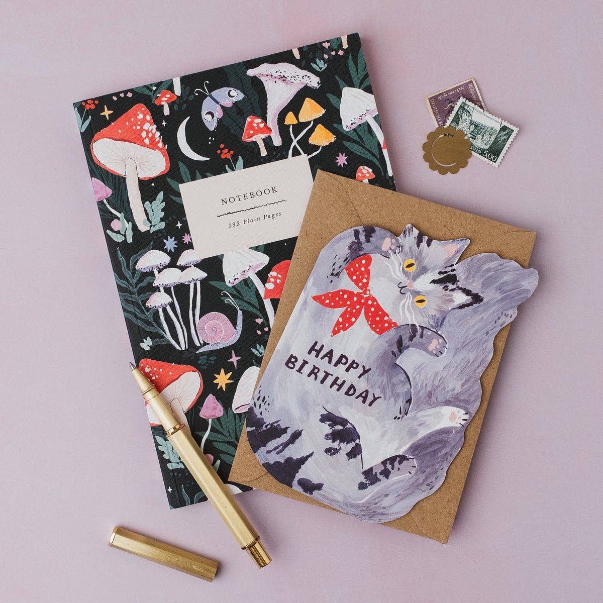 A styled flat-lay of stationery on a dusty pink background, featuring a black notebook with a colorful mushroom pattern, a brown greeting card with a gray cat illustration and 'HAPPY BIRTHDAY' text, a gold pen, and small postage stamps.