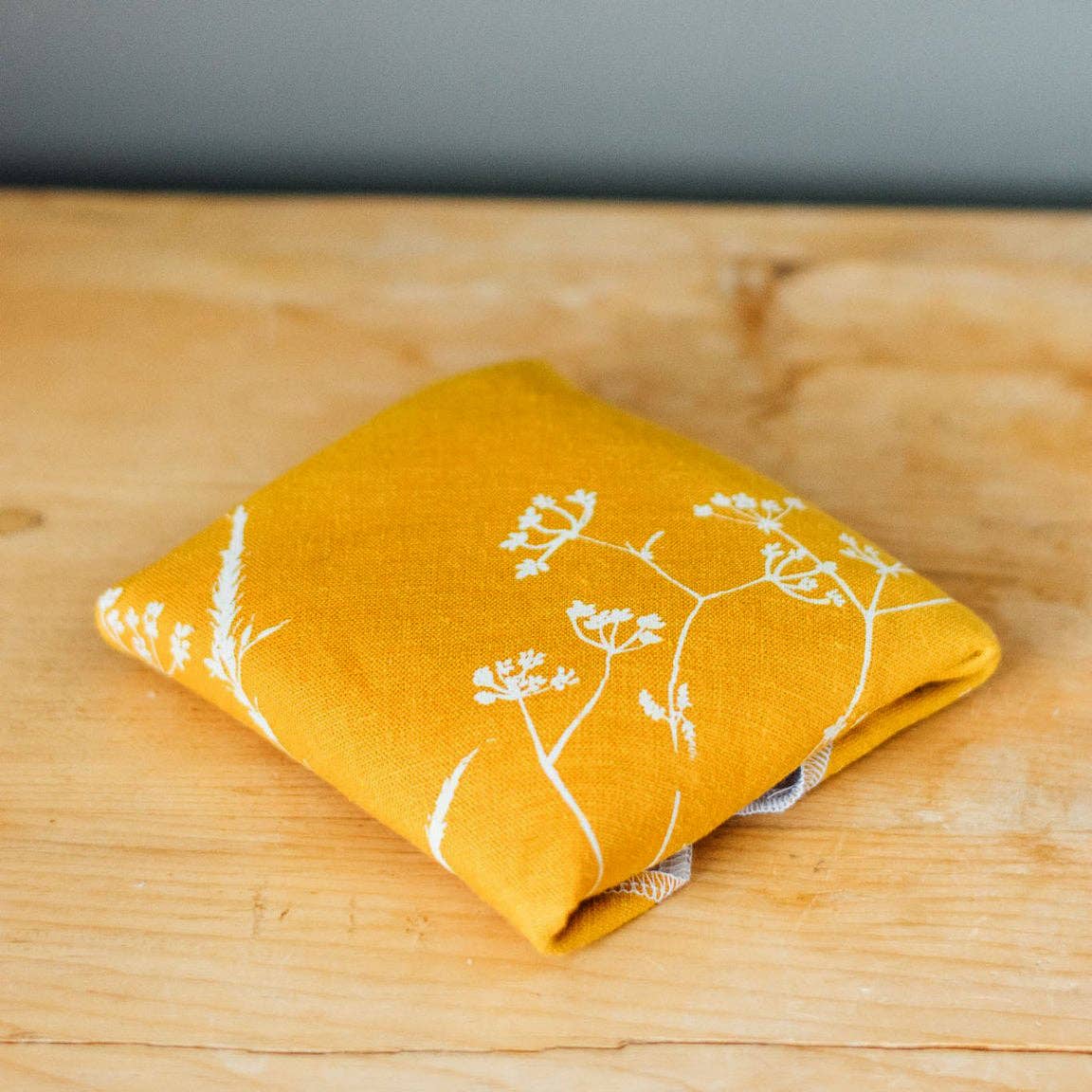 A folded mustard yellow linen cloth with a white botanical floral pattern, resting on a light wooden tabletop.