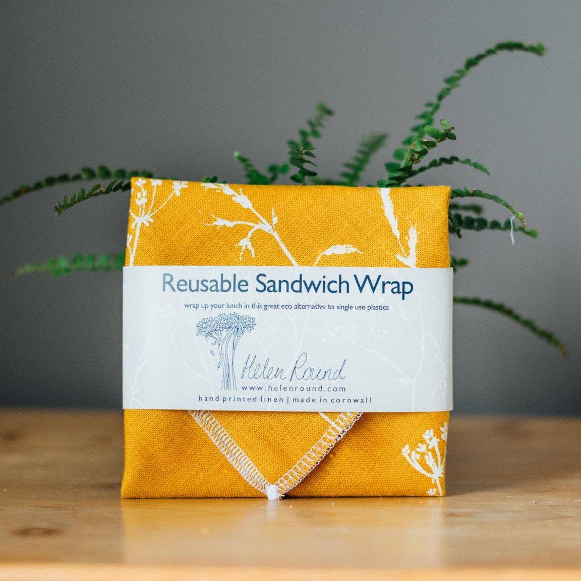 A folded reusable sandwich wrap in mustard yellow fabric with white botanical line drawings and a white paper band label reading 'Reusable Sandwich Wrap' and 'Helen Rand,' placed on a wooden surface with green plant stems against a grey background.