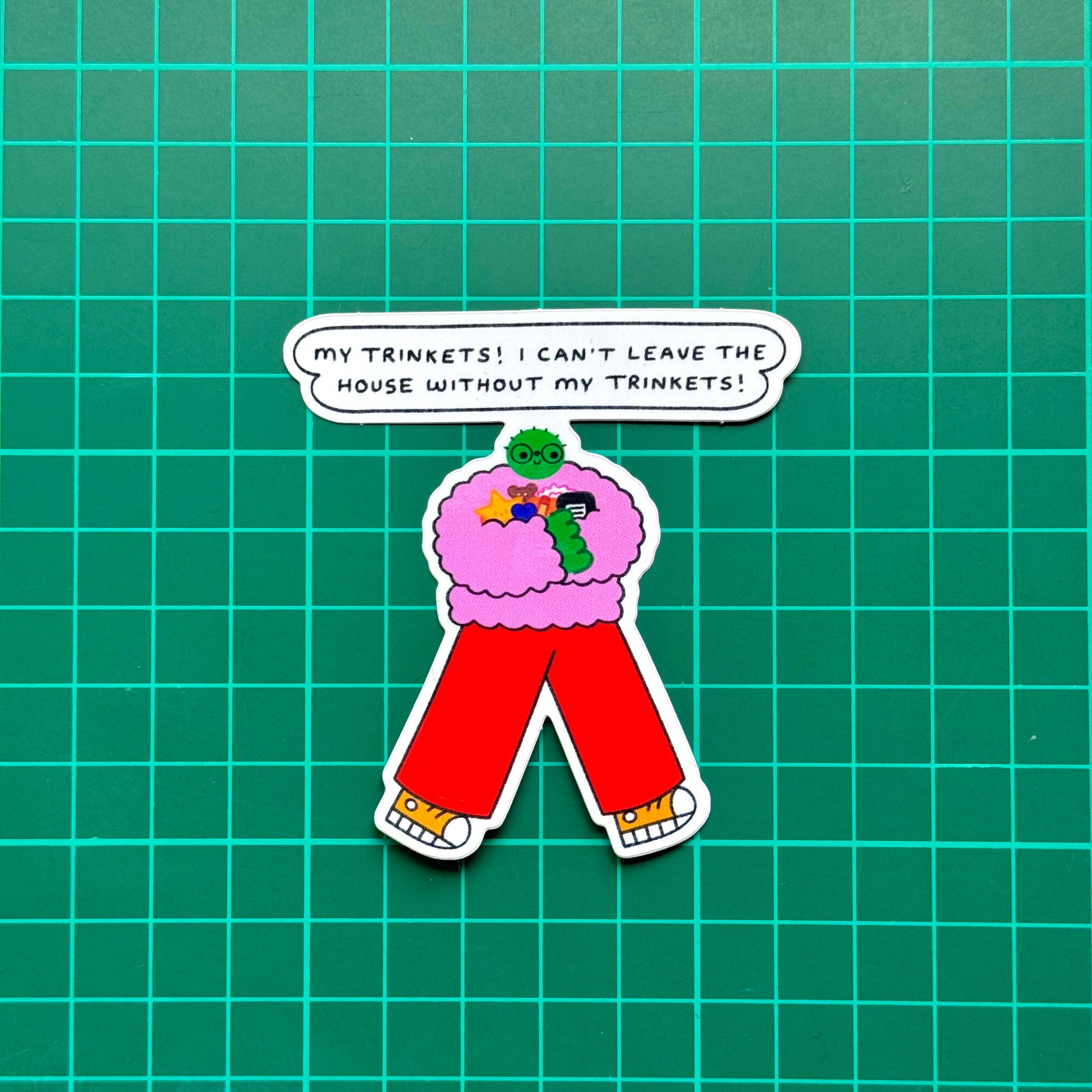 A sticker featuring a cartoon character with a pink fuzzy jumper, red pants, yellow shoes, and a green head, with a speech bubble above reading 'MY TRINKETS! I CAN'T LEAVE THE HOUSE WITHOUT MY TRINKETS!', on a green grid background.
