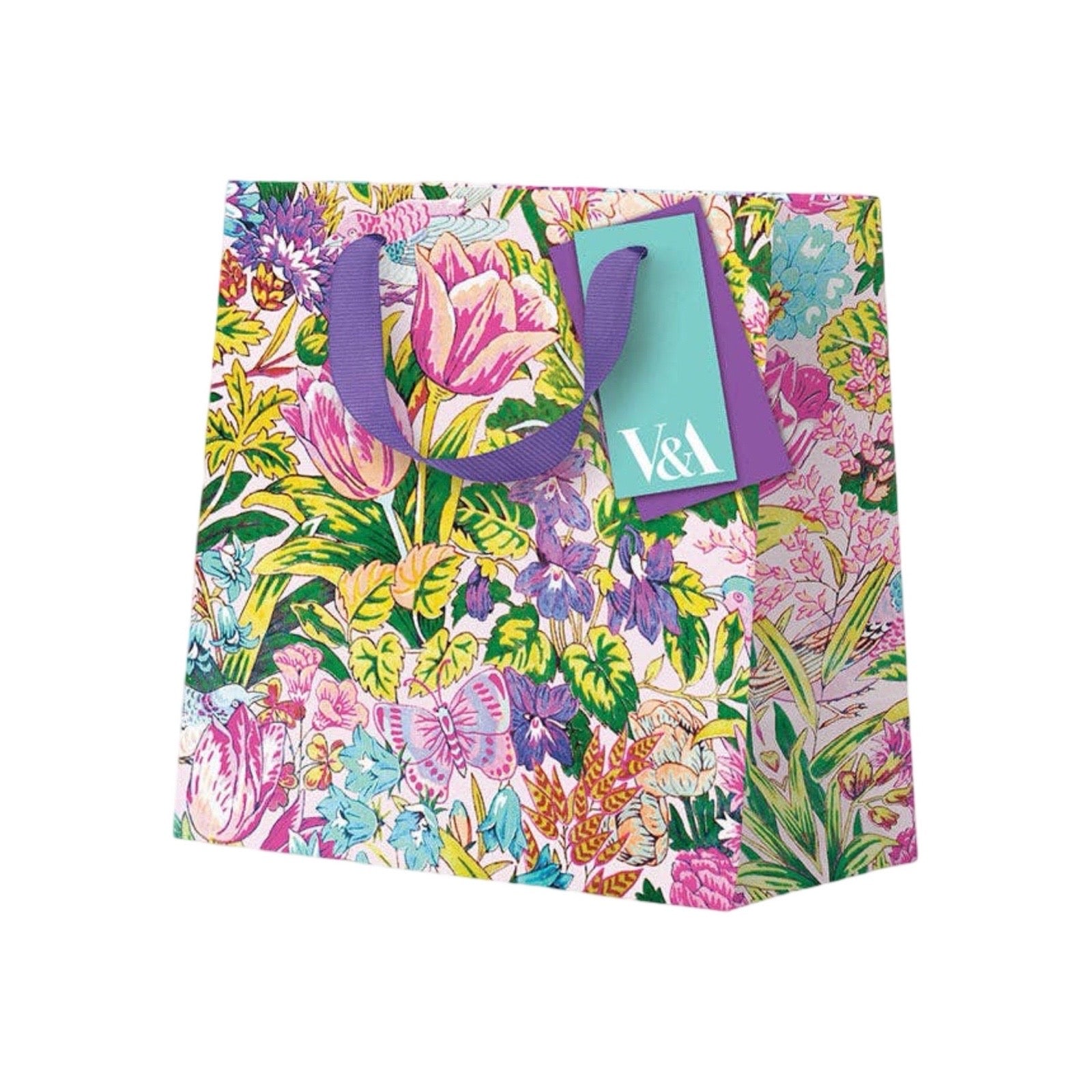 A colourful floral paper gift bag with purple handles and a turquoise tag featuring the white text 'V&A'. The bag is decorated with a dense, vibrant botanical pattern of pink, purple, green, and yellow flowers and leaves on a white background.