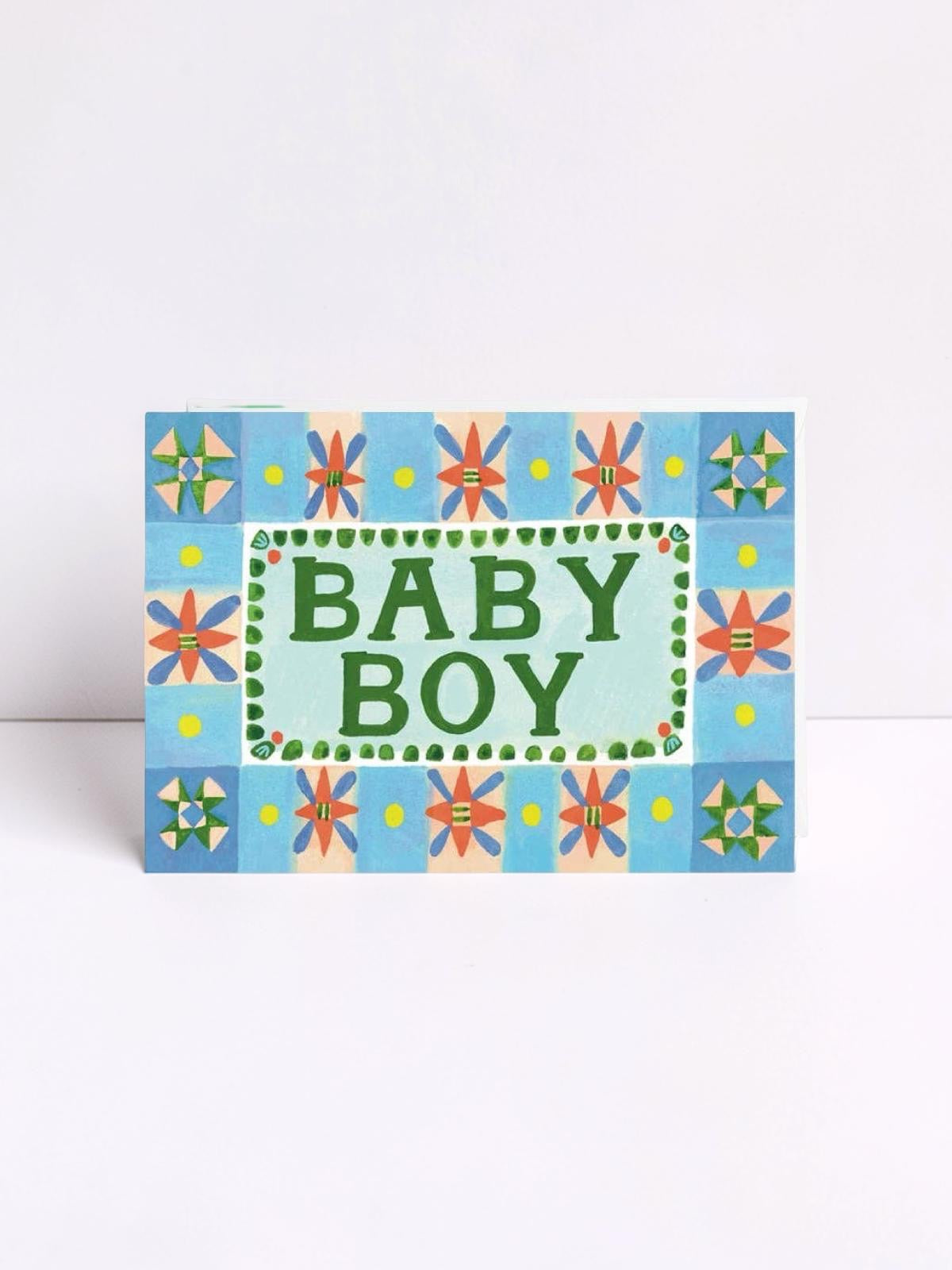 Card with 'BABY BOY' text on a blue floral patterned background, against a white wall.