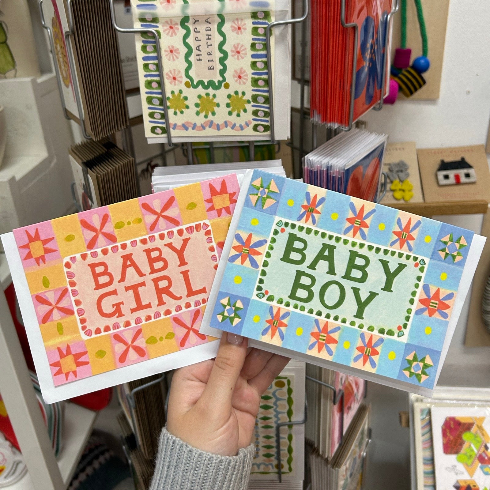 Hand holding two greeting cards, one for 'Baby Girl' and one for 'Baby Boy', with a store display in the background.