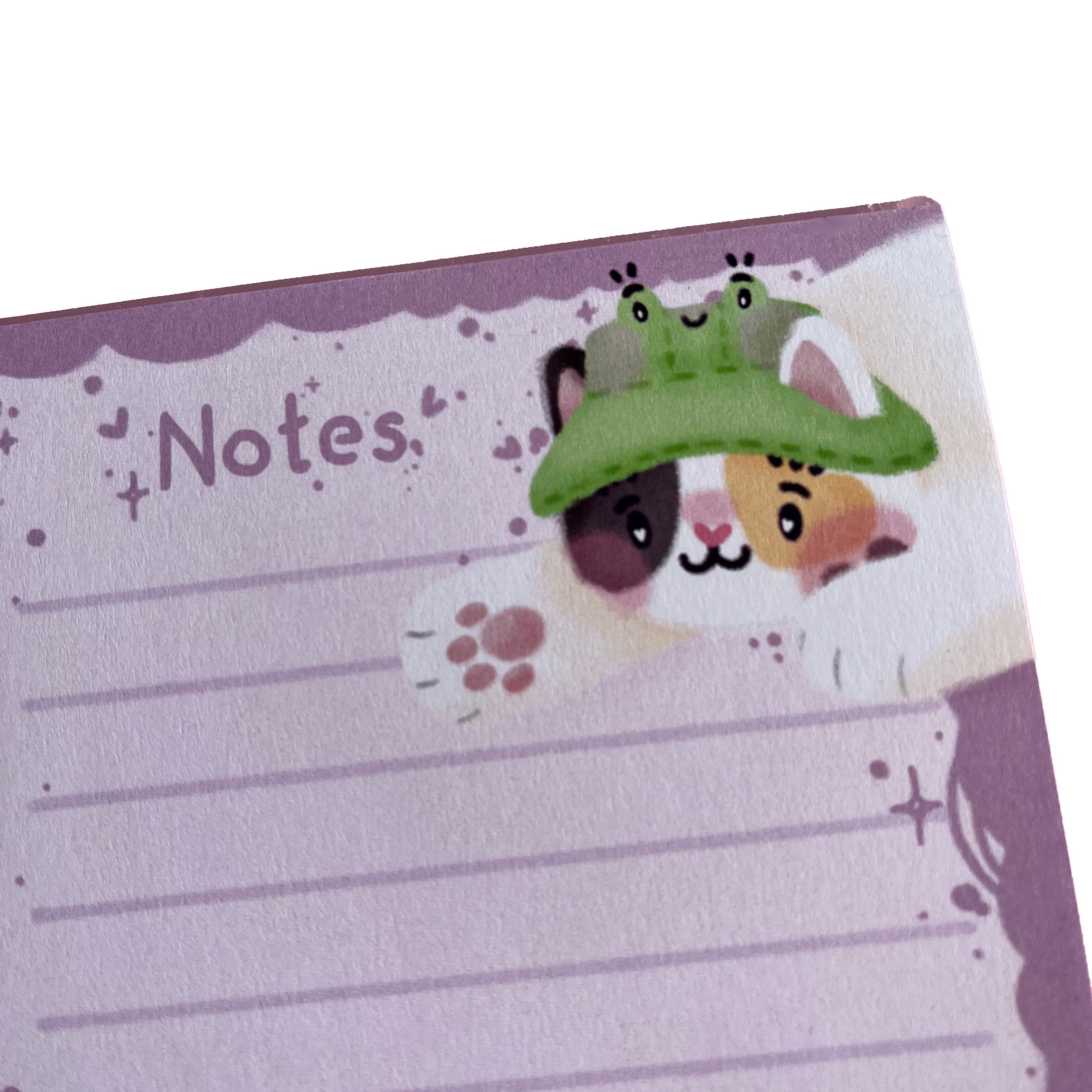 A 50-page pink tear-off lined notepad with Bucket the calico cat, wearing their frog bucket hat. Bucket is trying to catch your pen as you write your notes!