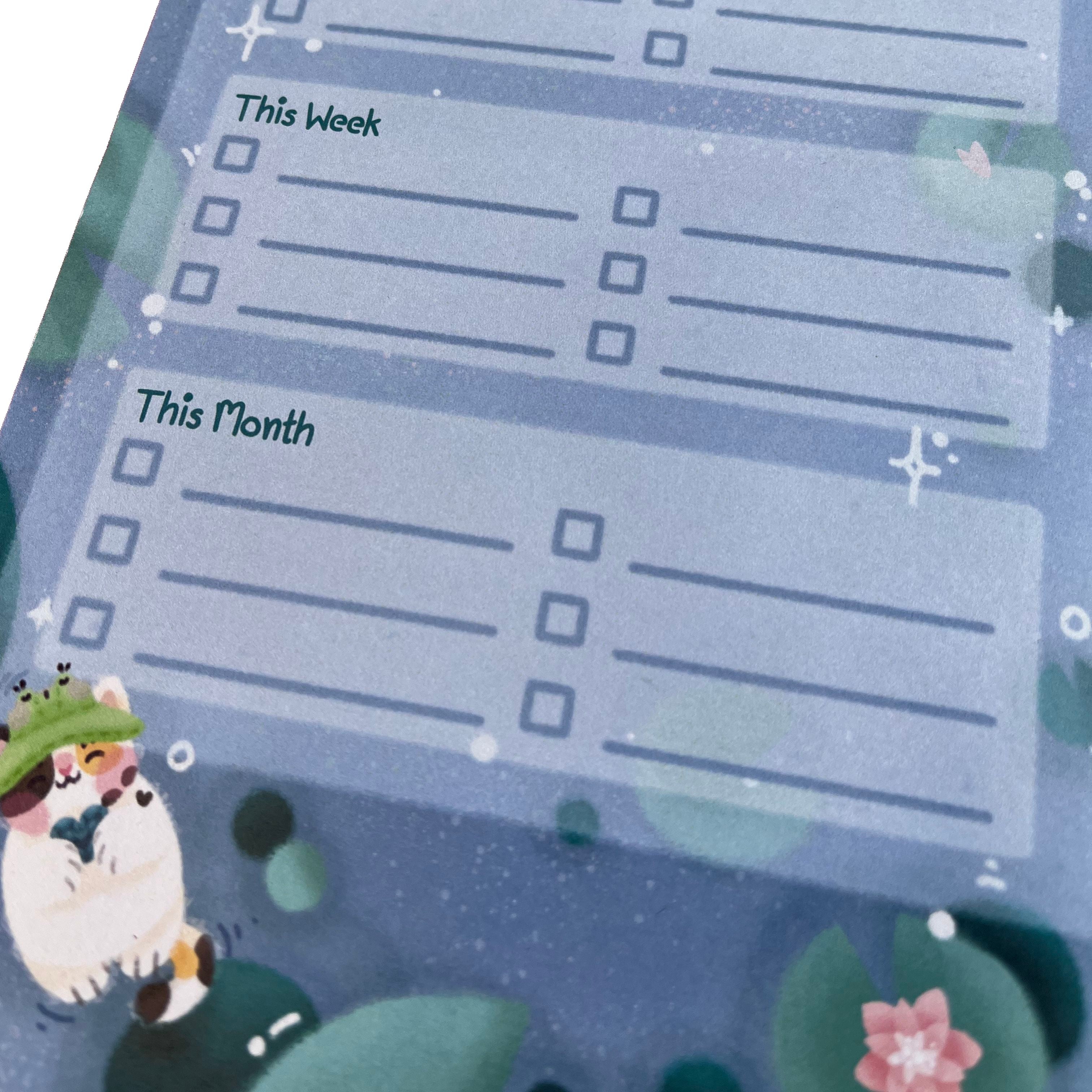 A 50-page tear-off lined notepad to help with your to do list items. With three sections to help prioritise tasks, labelled: 'today', 'this week', and 'this month', with check boxes. Text at top reads 'one thing at a time, not all at once'. The background features waterlily pads and a calico cat swimming on it's back while holding a heart shaped rock.&nbsp;