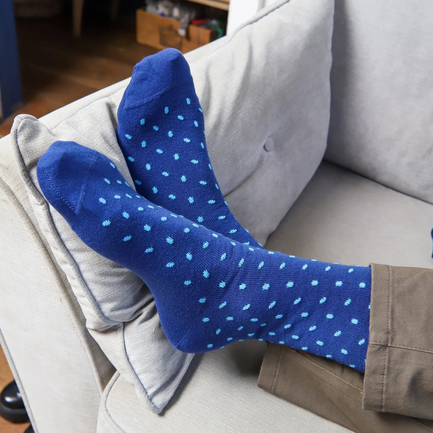 A pair of blue crew socks with light blue polka dots worn on feet resting on a light gray couch cushion.