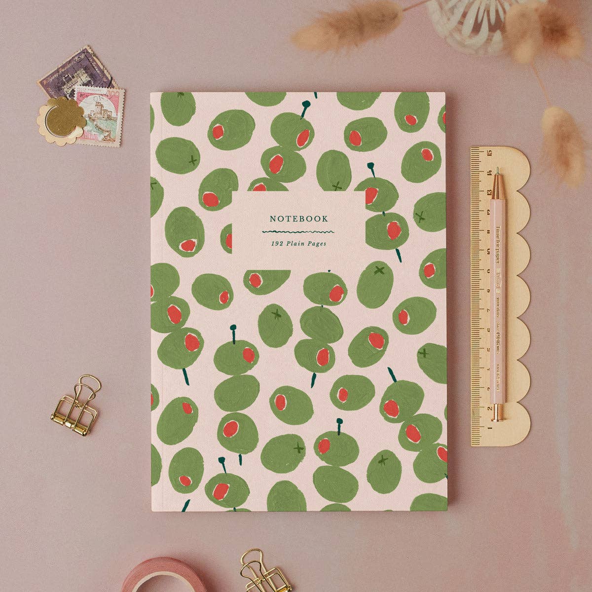 Notebook with green olive pattern on a pink background, surrounded by stationary items and a plant.