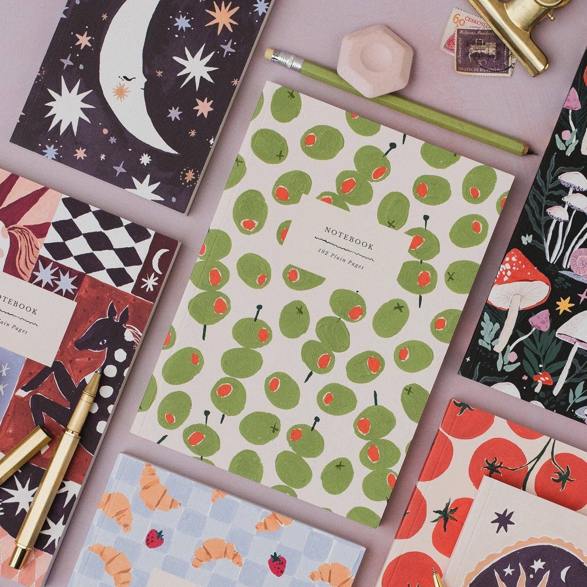 A styled flat-lay of patterned notebooks on a dusty pink background, with a central cream notebook featuring a green avocado print and red seed details. Surrounding notebooks include celestial, checkered, mushroom, strawberry, and croissant motifs, accented with gold and pink pencils and a pink geometric pen stand.