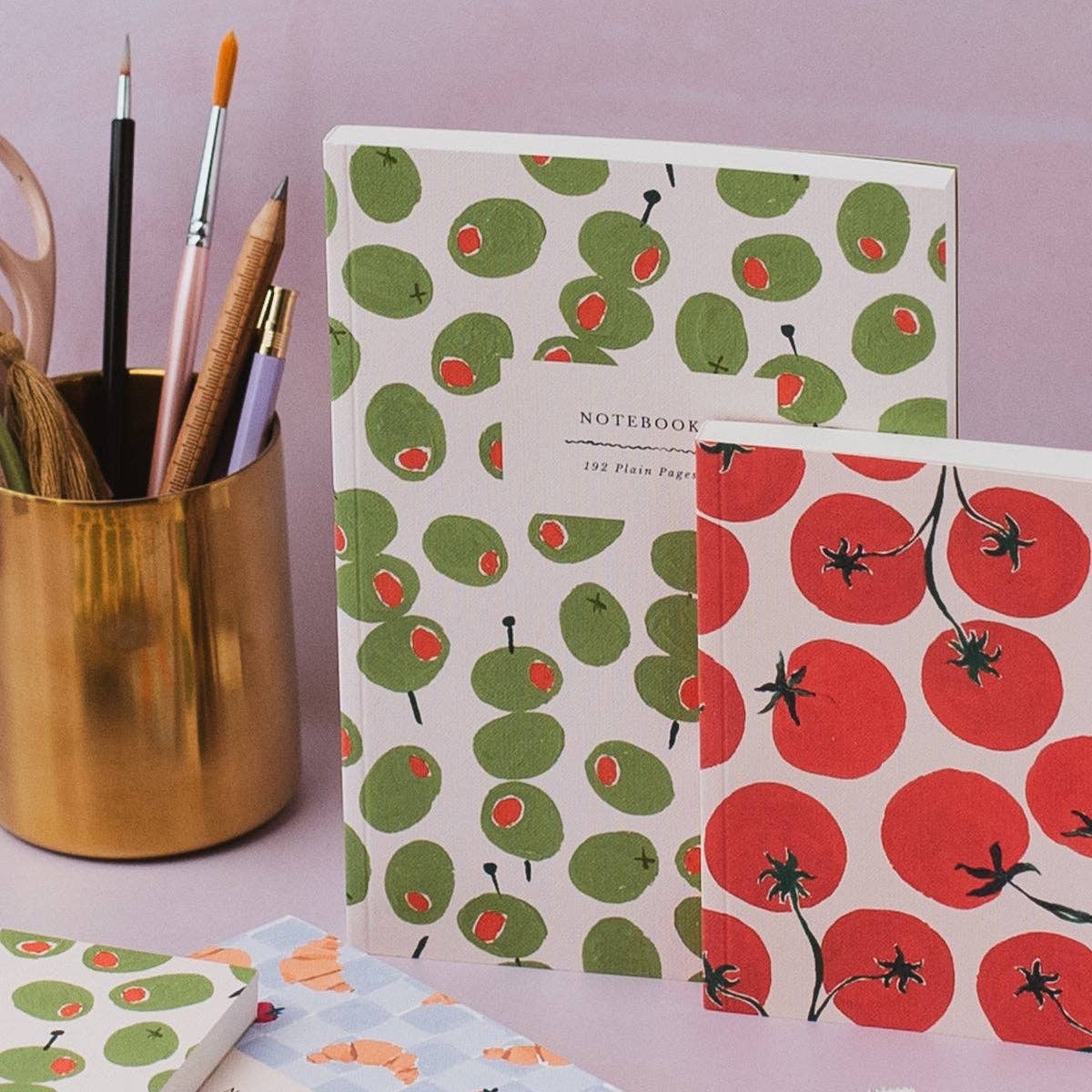 A set of patterned notebooks displayed on a lavender surface next to a gold cup holding paintbrushes and pencils. The notebooks feature retro illustrations of green olives with red centers and red tomatoes with black stems.