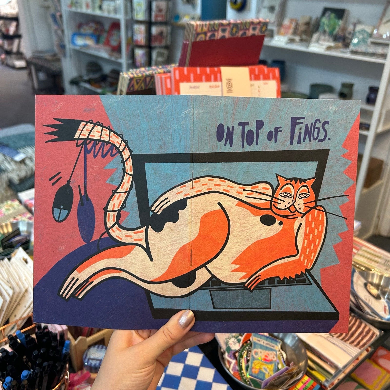 A hand holds a notebook featuring a stylized orange and cream cat lounging on a laptop with the text 'ON TOP OF FINGS.' The background shows a busy retail store shelf with various items.