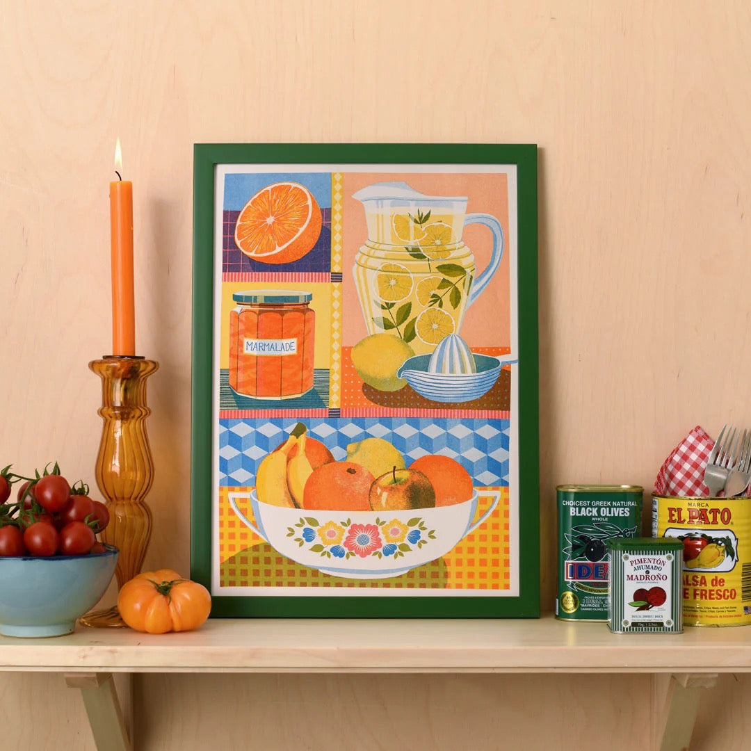 Framed colorful artwork of a kitchen scene with a pitcher, lemons, and oranges on a beige wall.