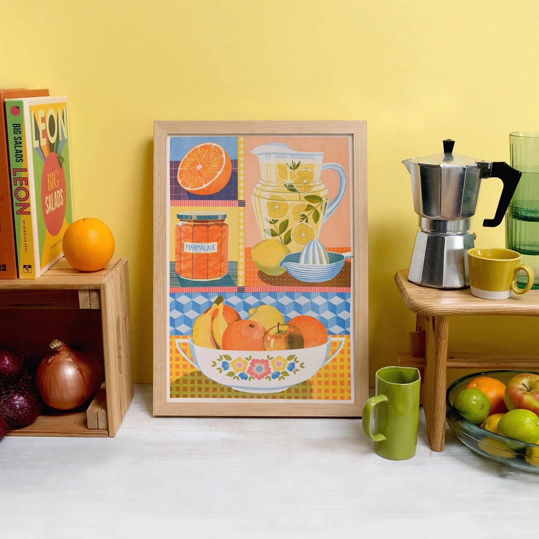 Framed artwork of a colourful still life scene with fruits and kitchen items on a yellow wall.