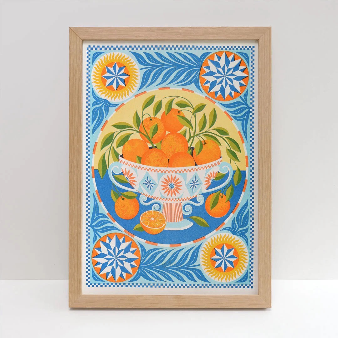 Framed art work of a decorative bowl of oranges on a blue background, against a white wall.