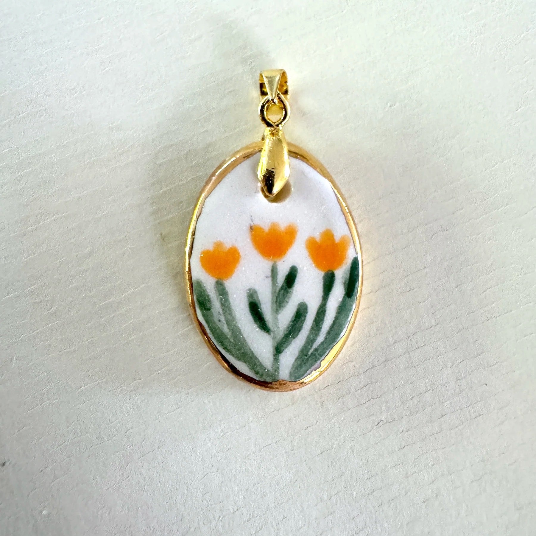 Gold pendant with a ceramic design of orange flowers on a white background.