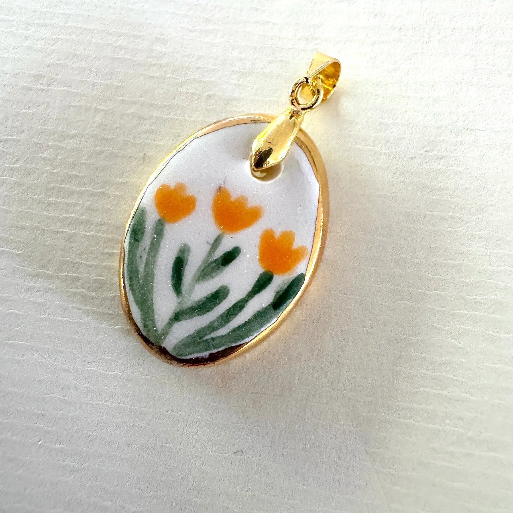 Gold pendant with a ceramic disc featuring orange flowers and green leaves on a white background.