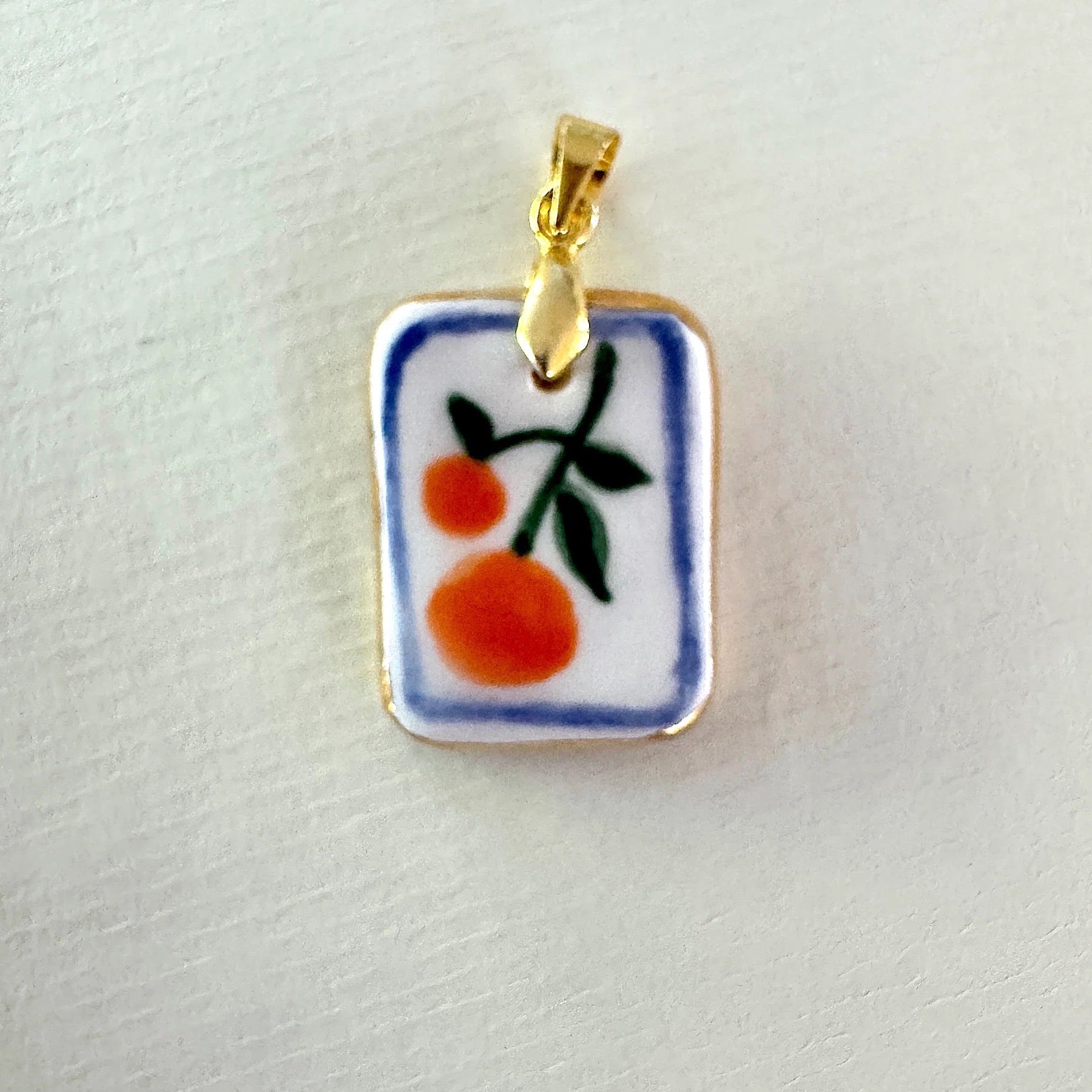 Small rectangular pendant with a ceramic design of oranges on a gold chain against a light gray background.