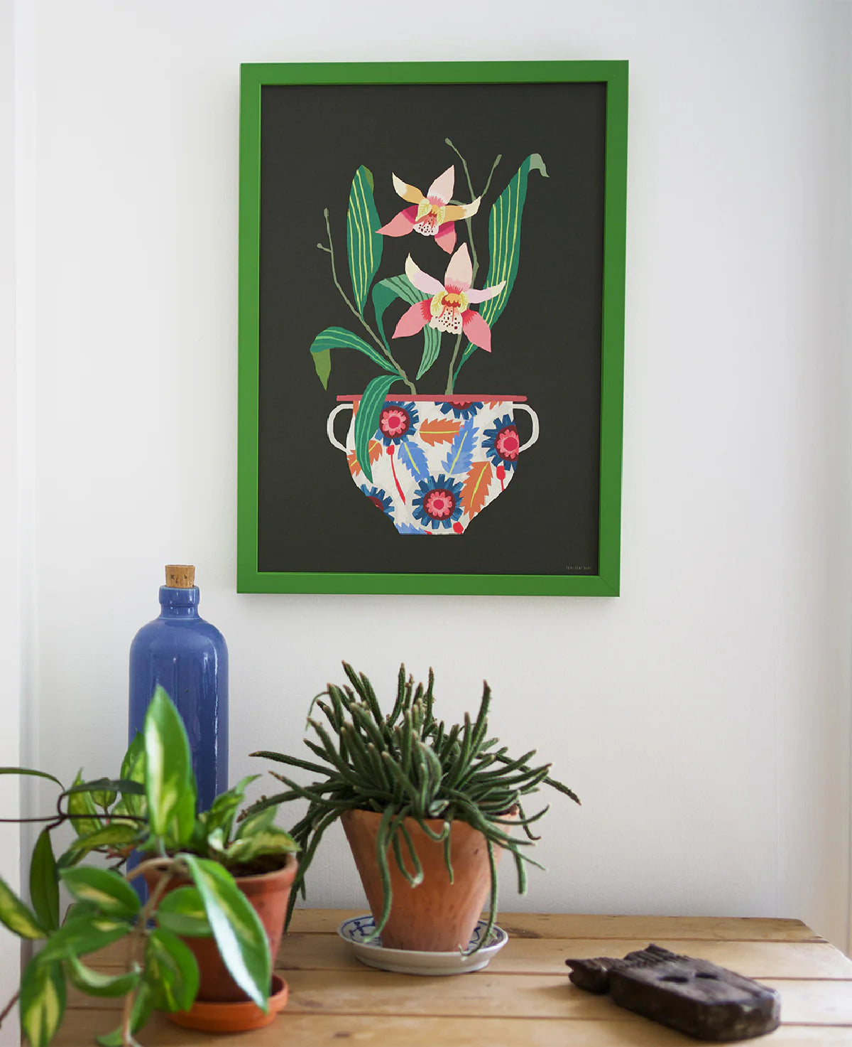 Framed floral artwork on a wall with potted plants below.