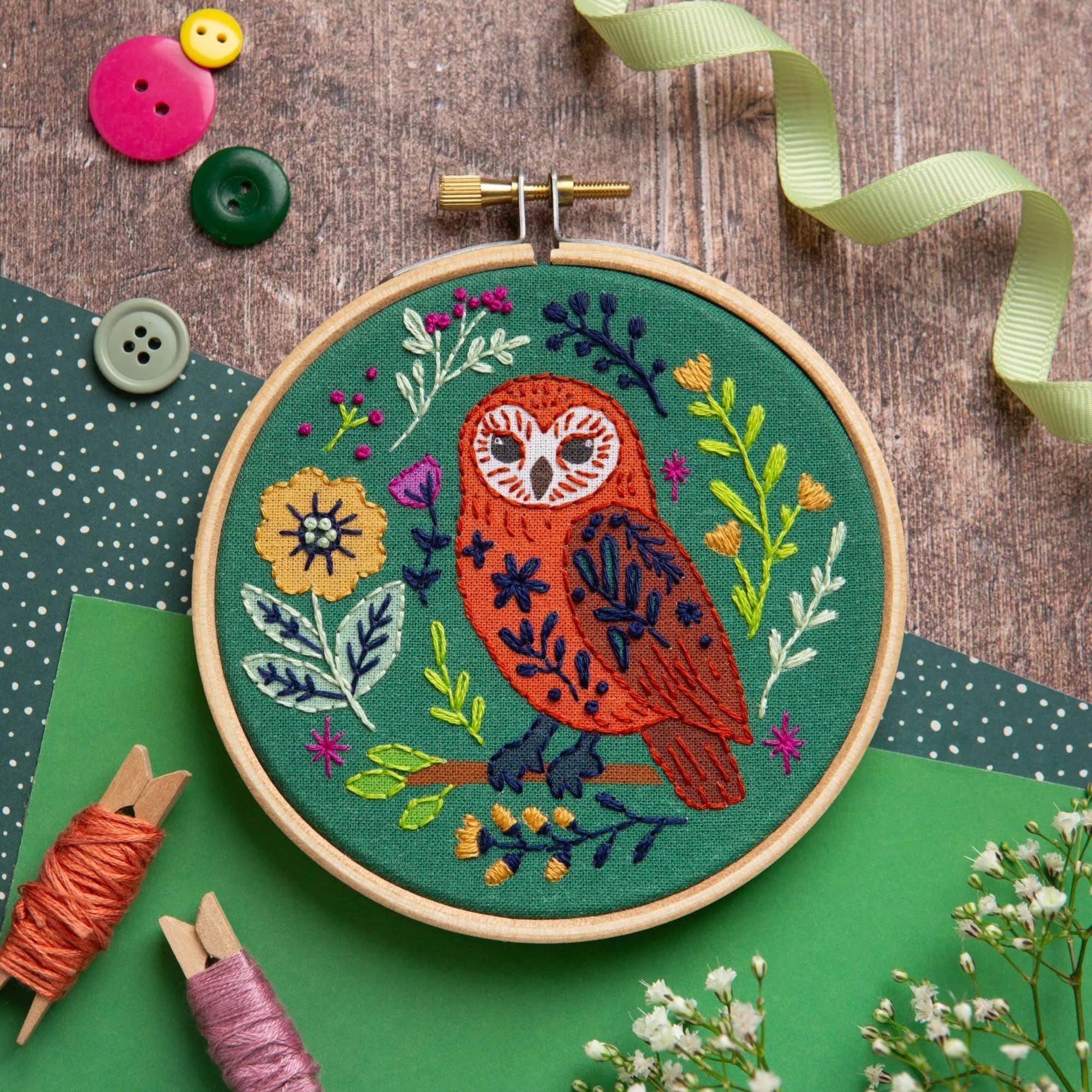 A round embroidery hoop holding a completed floral owl embroidery on green fabric, styled on a wooden surface with buttons, ribbon, spools of thread, and small white flowers.