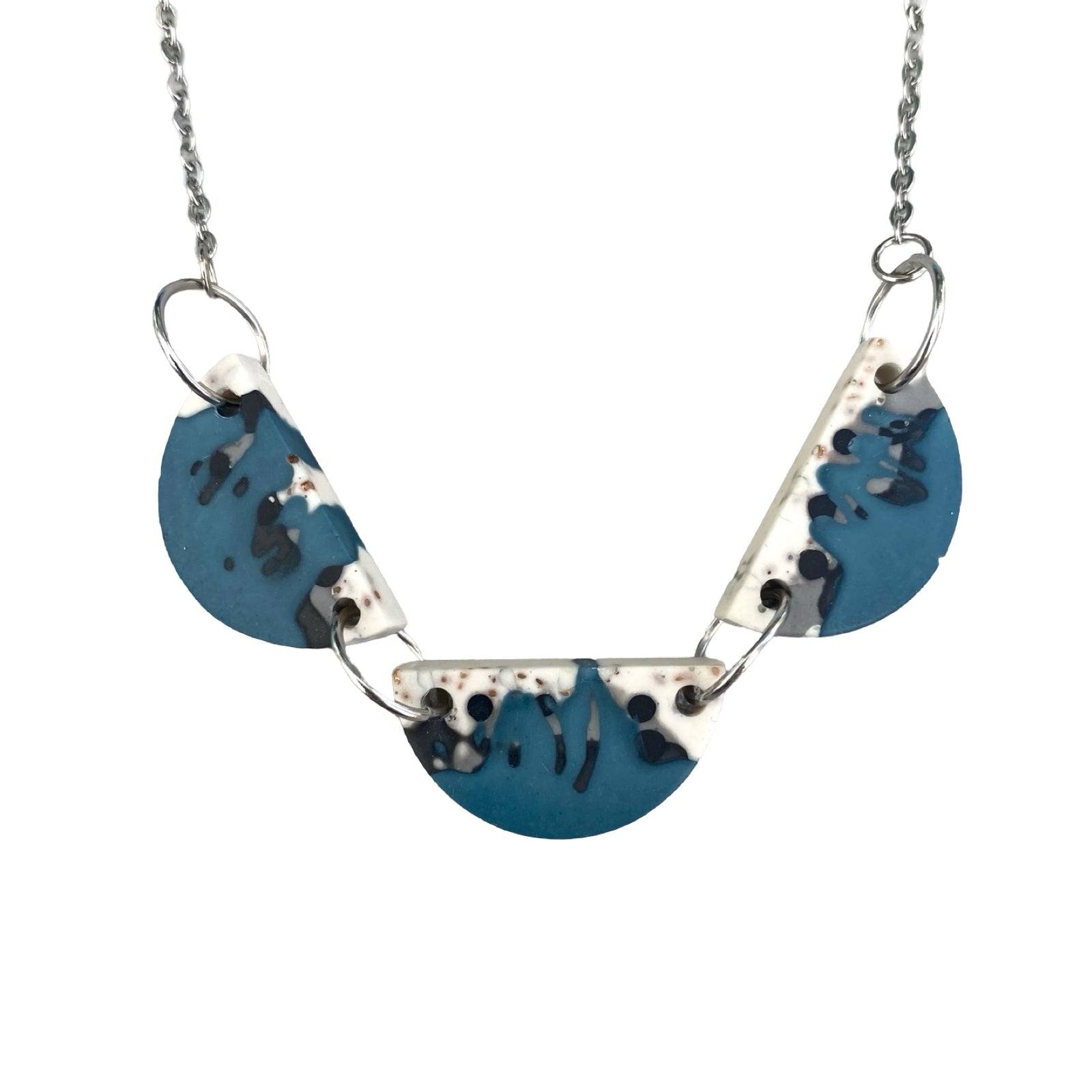 A necklace with three half moon resin shapes in blue with a black and white paint design held together with silver findings on a silver chain.