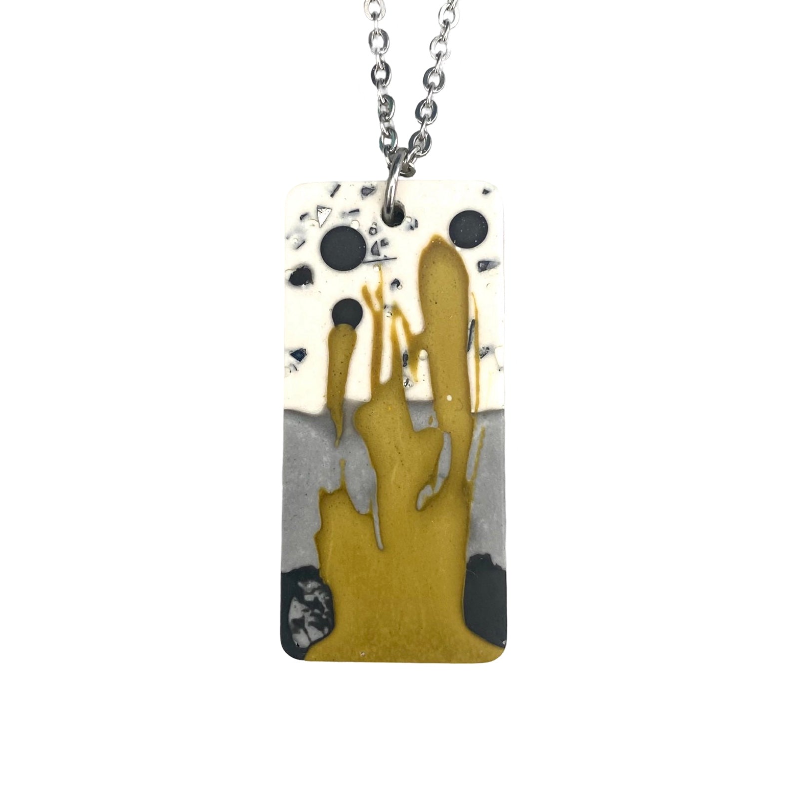 A rectangular pendant necklace iwith a muted paint design in mustard, black, grey and white on a silver chain.